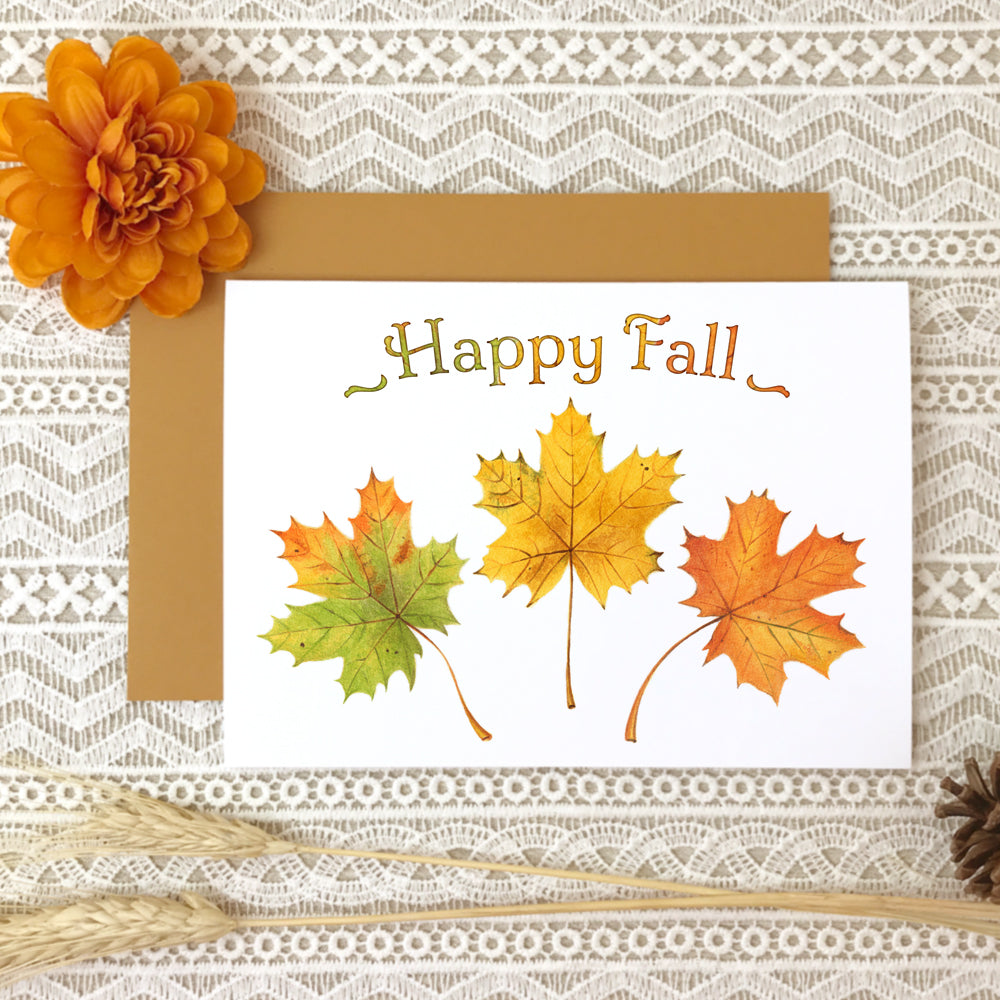Autumn greeting card titled "Happy Fall" with watercolor paintings of three maple leaves in green, yellow and orange.